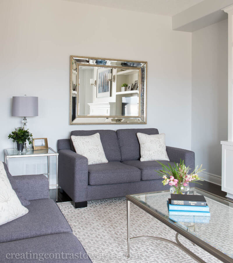 Transitional Living Room With Grey Loveseat With White Accent Cushions And Large Bevelled Mirror And Glass Coffee Table And Patterned Area Rug