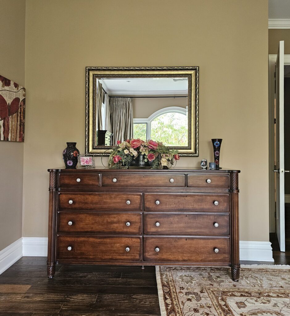Before photo of dark wood dresser with floral arrangement and ornate gold mirror on beige wall, principal bedroom redesign by clairejefford in Burlington, Ontario.