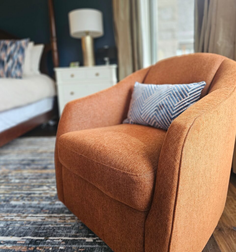 Detail of rust orange custom swivel chair with blue and white lumbar pillow on patterned rug, moody bedroom refresh by clairejefford interior design Burlington.