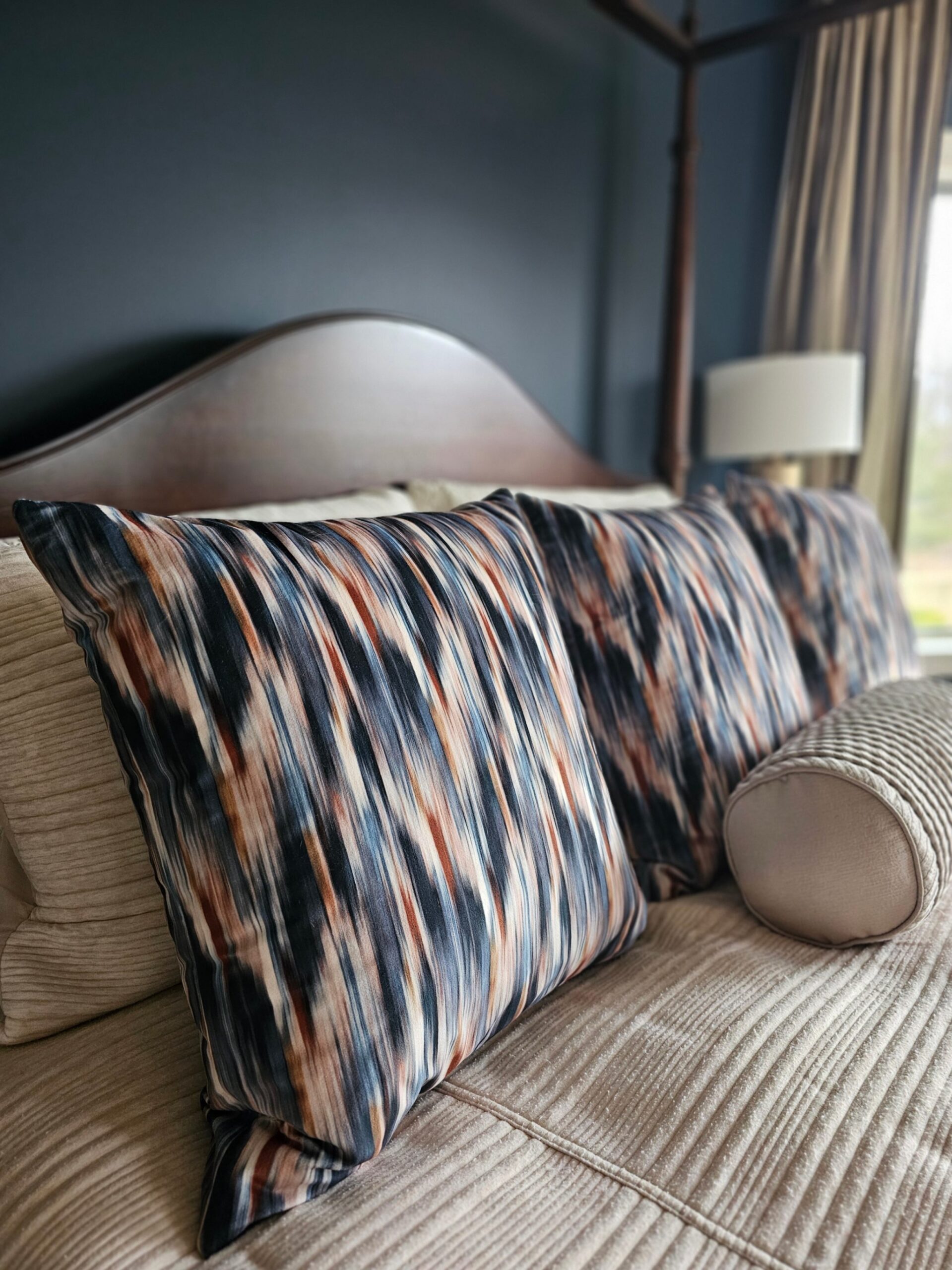 Close-up of custom velvet accent pillows in rust, navy, and cream pattern on cream bedding, styled against navy wall and dark wood four-poster bed by clairejefford.